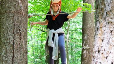 Child wearing a monkey hat balances on a rope between trees in a lush forest.