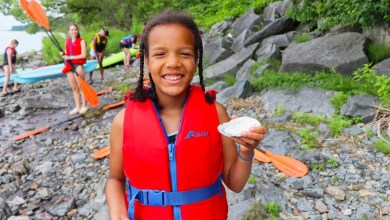 Smiling girl in a red life vest on a rocky shoreline, holding a shell toward the camera, with kayaks and helpers in the background.