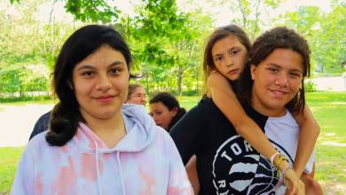 Group of four teen friends in a sunny park; two girls in front smile, while one girl hugs another from behind.