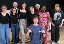 Group of ten people posing together on a stage, smiling at the camera, casual attire
