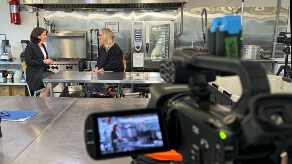 Two women in a stainless steel kitchen interview set chatting at a counter while a video camera films them in the foreground.
