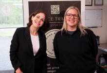 Two women pose for a photo indoors in front of a banner featuring a QR code.
