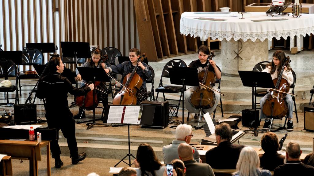 Conductor leads a small string ensemble of cellists on a church stage with an attentive audience in the foreground.