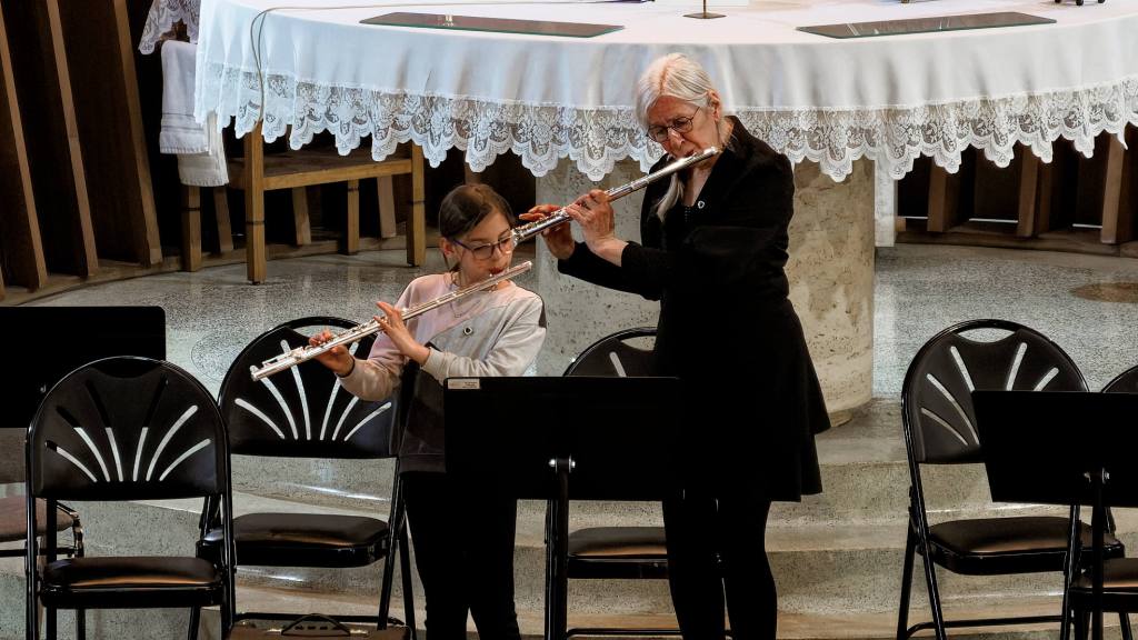Adult woman teaches flute to girl student in a church hall with a lace-covered table and empty chairs nearby.