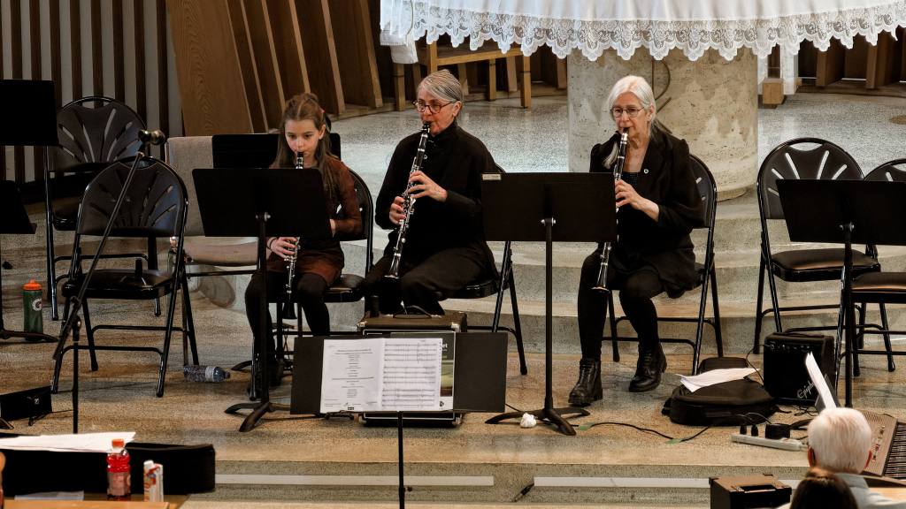 Three musicians—a young girl and two older women—playing clarinets in a church hall with music stands and chairs nearby.