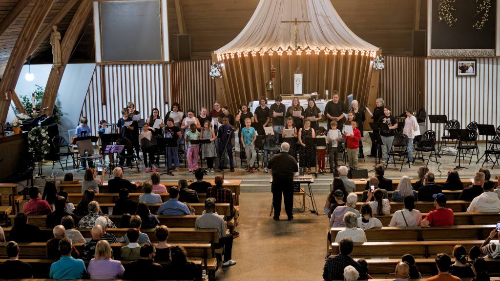 A large group of children and adults sing on a church stage while a conductor stands in front, with an audience in pews watching.