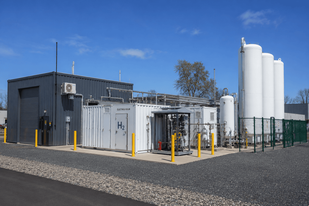 Industrial hydrogen storage site with a white H2 container, piping, and tall white storage tanks under a blue sky.
