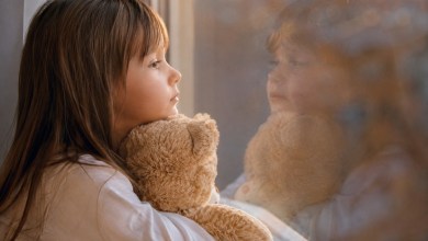 Young girl gazing out a window while hugging a beige teddy bear, her reflection visible on the glass.