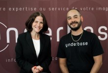 Two people pose for a photo in front of a burgundy backdrop with 'underbase' branding; woman in a black blazer and white top smiles beside a man in a black 'underbase' shirt and chain necklace.
