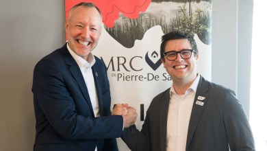 Two professionally dressed men shake hands and smile in front of a banner.