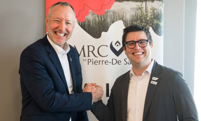 Two professionally dressed men shake hands and smile in front of a banner.