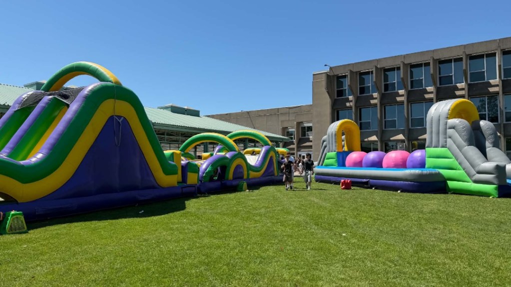 Colorful inflatable obstacle course set up on a grassy field beside a modern building; several people stand nearby on a sunny day.
