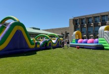 Colorful inflatable obstacle course set up on a grassy field beside a modern building; several people stand nearby on a sunny day.