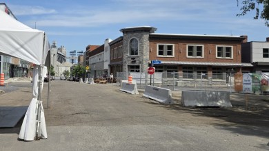 A daytime construction street with barricades, cones, and a brick building; tents set up on the left and a stop sign reading ARRET in the foreground.