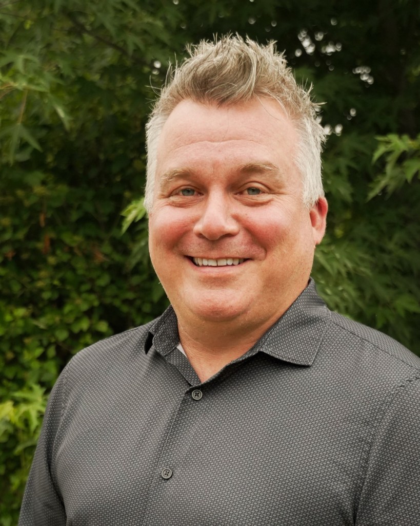 Portrait of a smiling man with short gray hair outdoors, wearing a dark patterned button-up shirt, with green foliage in the background, looking at the camera.