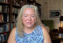Woman with blonde, curly hair sits in a cozy home library, smiling with bookshelves behind her.
