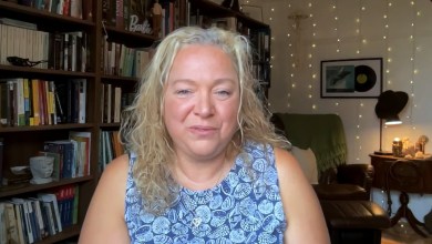 Woman with blonde, curly hair sits in a cozy home library, smiling with bookshelves behind her.