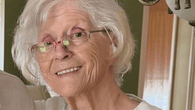 Smiling elderly woman with white hair and pink glasses indoors near a chandelier in a home setting