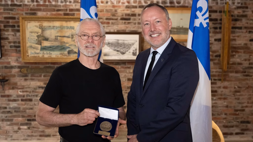 Two men pose after a medal award: the man in a black shirt holds an open presentation box with a medal while the man in a navy suit smiles beside him, flags and a brick wall backdrop behind them.