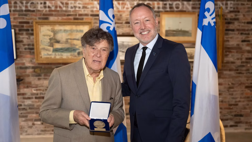 Two men in suits smile for the camera; the older man holds an open medal case displaying a bronze award during a ceremony with flags in the background.