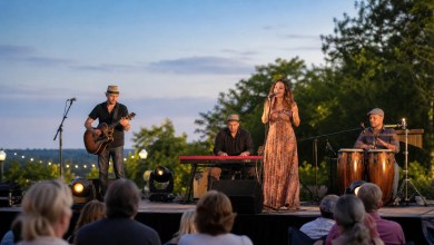 Outdoor musical performance at dusk with a female singer in a long patterned dress accompanied by a guitarist, keyboardist, and conga players on stage, audience in foreground.